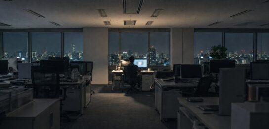 照明の落ちた深夜のオフィスで一席だけ灯るデスク